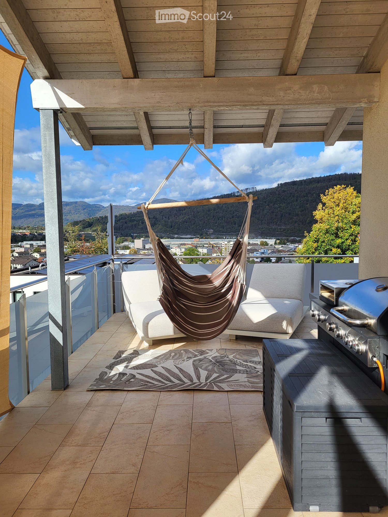 Modern balcony with a view of mountains, featuring a white sofa, a hanging hamaca, a patterned rug, and a grill.
