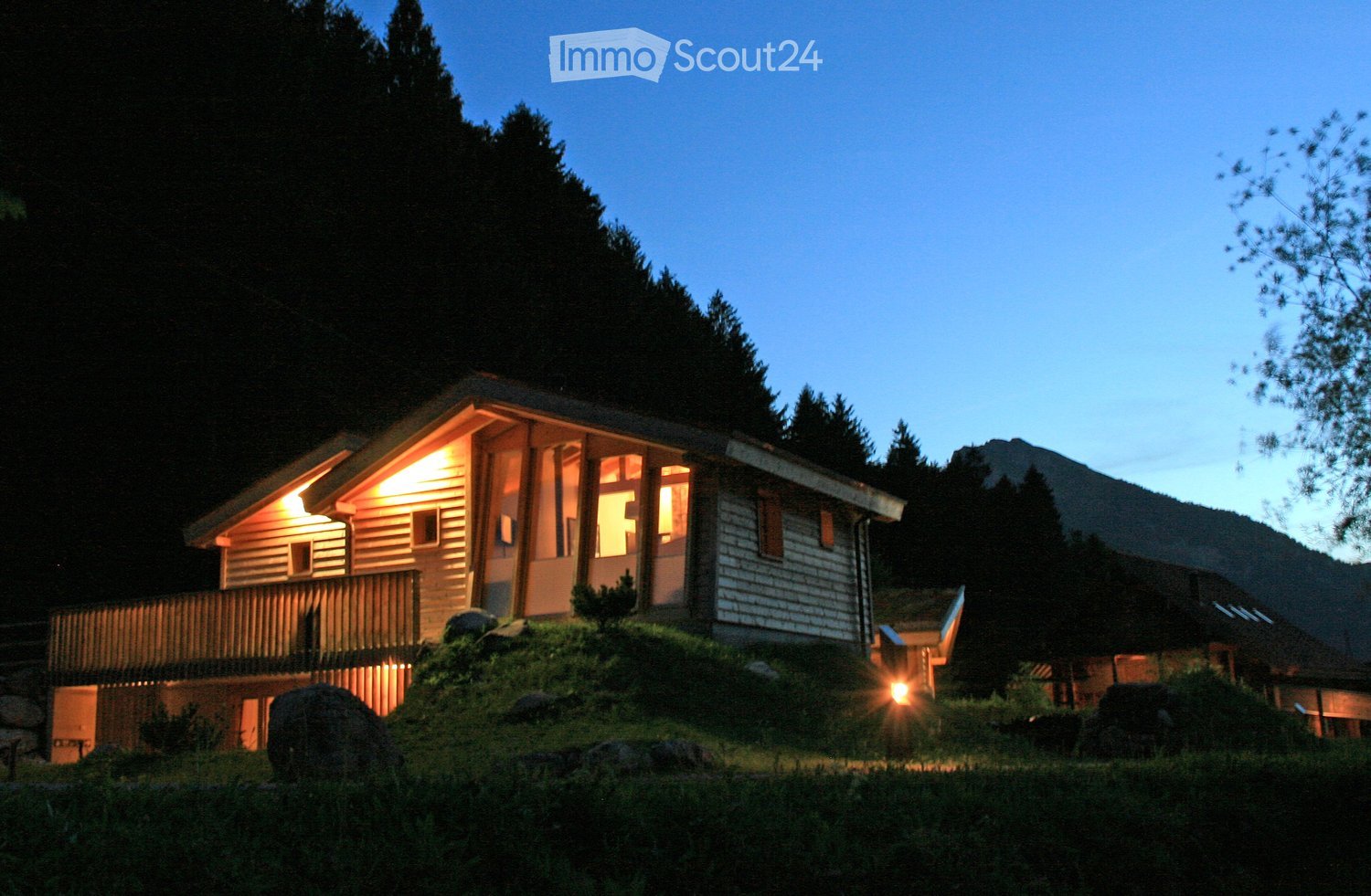 A cozy wooden house with a raised ground floor, illuminated windows, and a small balcony, surrounded by trees and a grassy area.