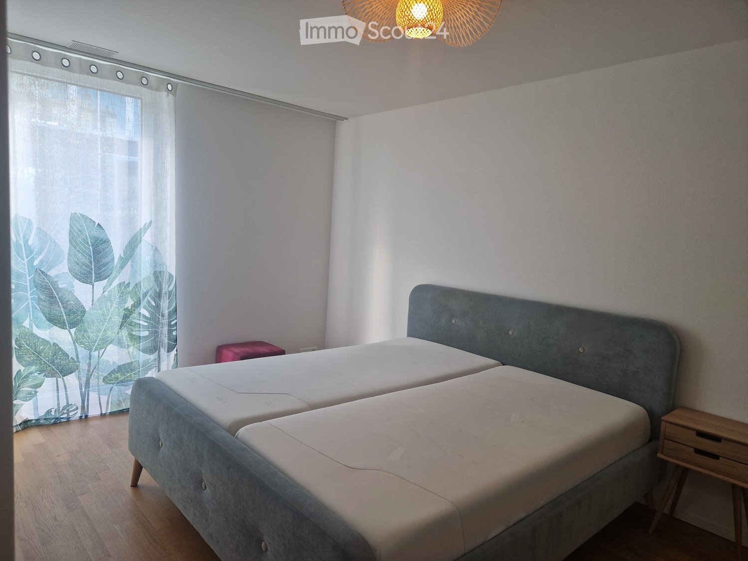 Modern bedroom with two single beds, grey headboard, wooden nightstand, and large window with leafy curtain.