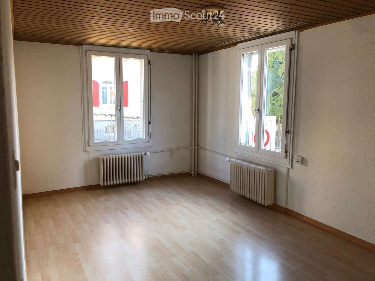 Empty room with wooden flooring, white walls, and two radiators. Features large windows with white frames and a wooden ceiling with recessed lighting.