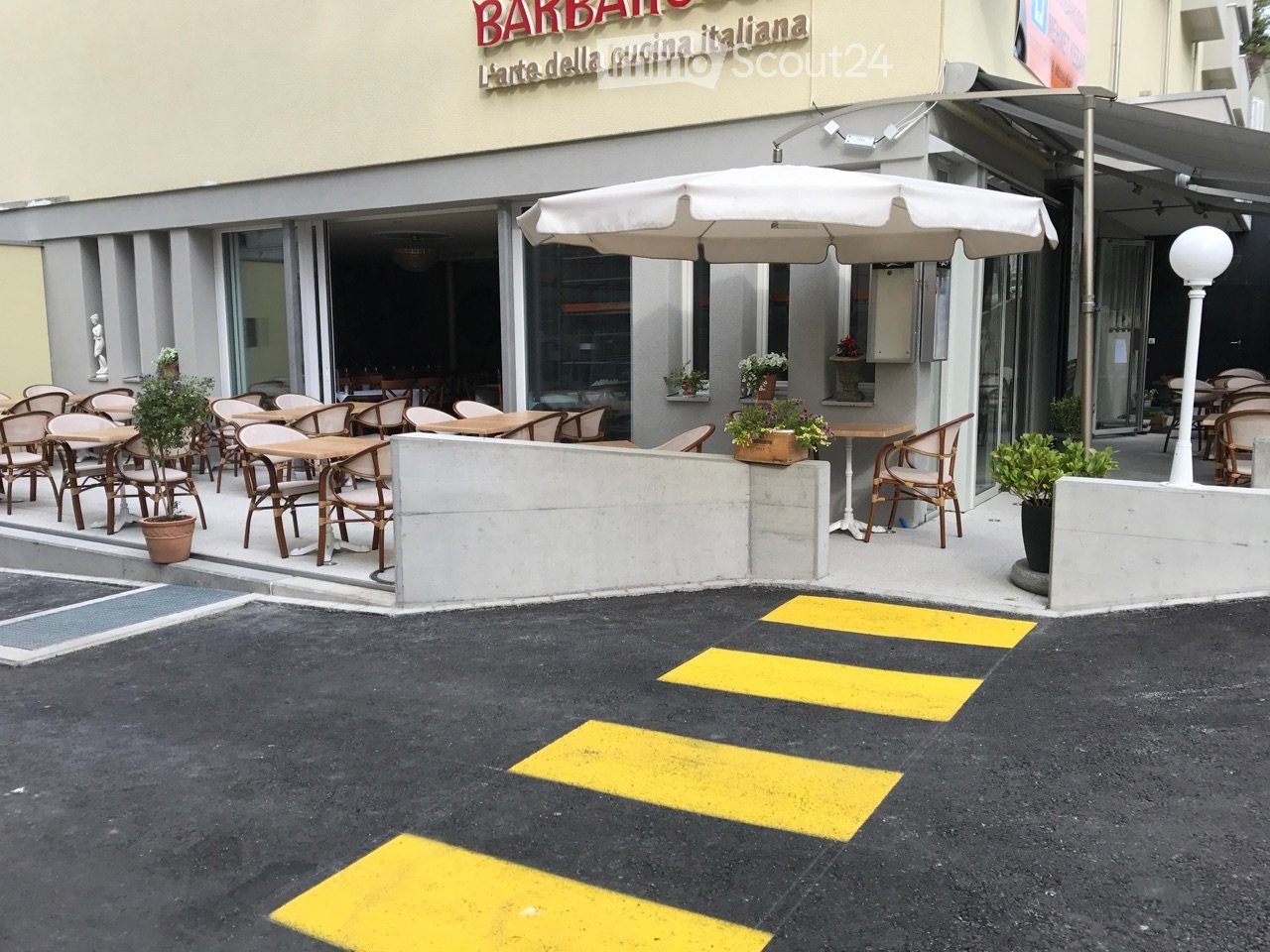 Outdoor seating area of a restaurant with tables, chairs, and an umbrella, surrounded by potted plants and a building entrance.