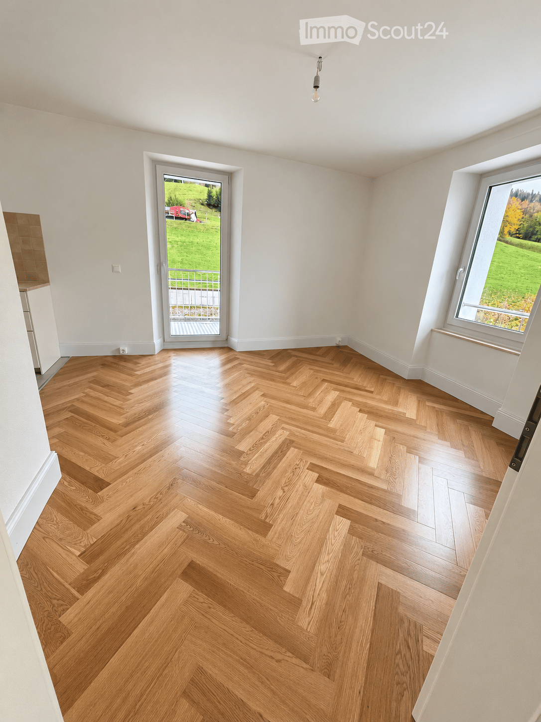 Empty room with herringbone-patterned wooden flooring, white walls, large windows, and a sliding glass door leading to a green outdoor area.
