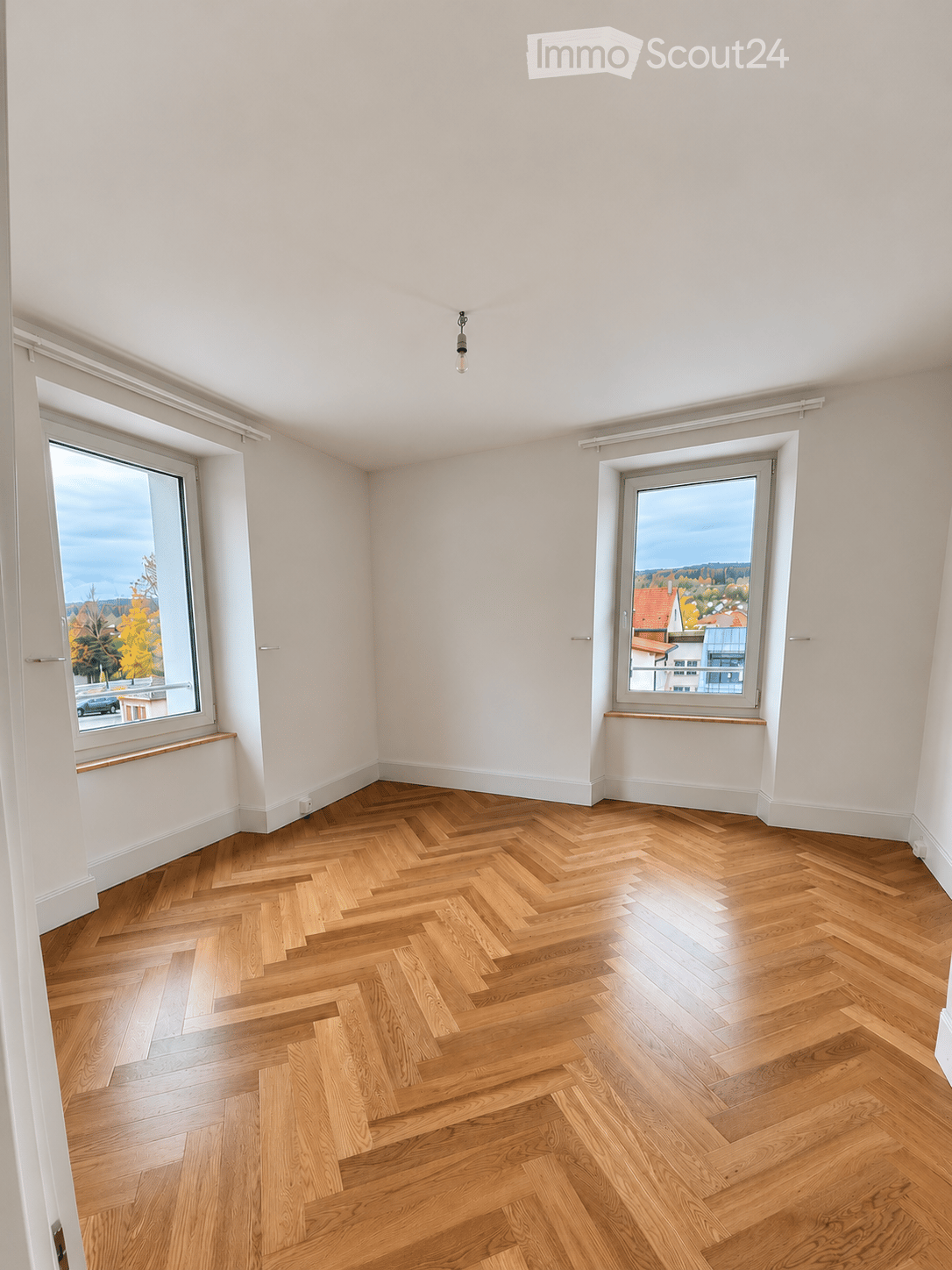 Empty room with light-colored walls, herringbone wooden flooring, and large windows offering a view of the outside. No furniture or equipment is present.