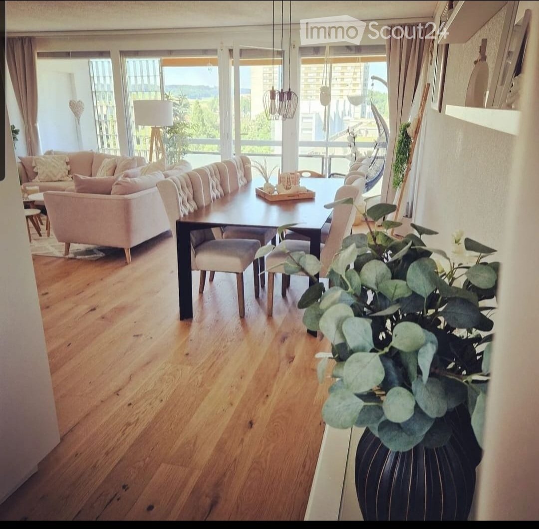 Open-plan living and dining area with wooden flooring, large windows offering a scenic view, a cream sofa, a dark wooden dining table with chairs, and a decorative flower vase on a side table.