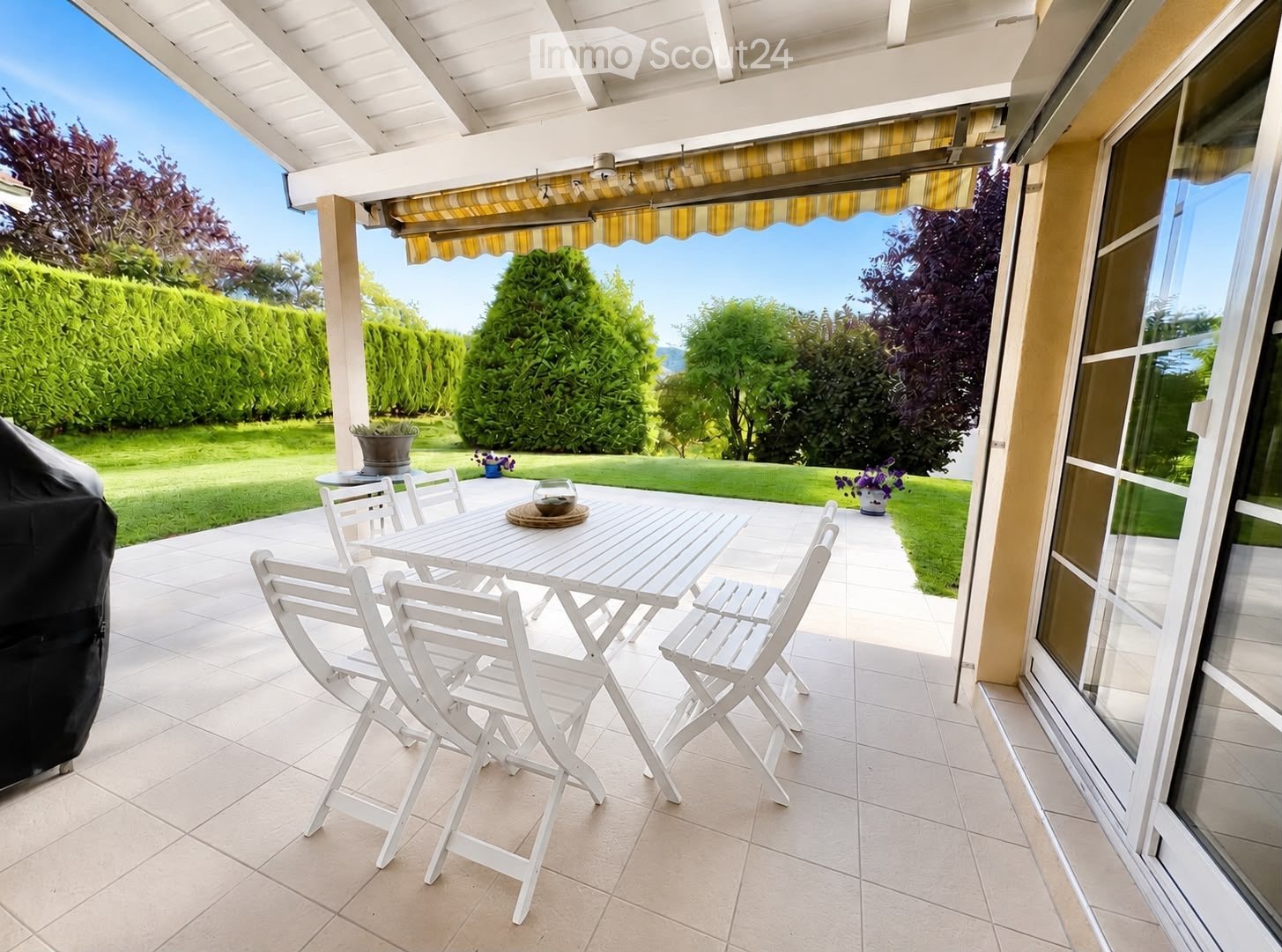 A covered patio with a white table and chairs, overlooking a well-maintained garden with green hedges and trees.