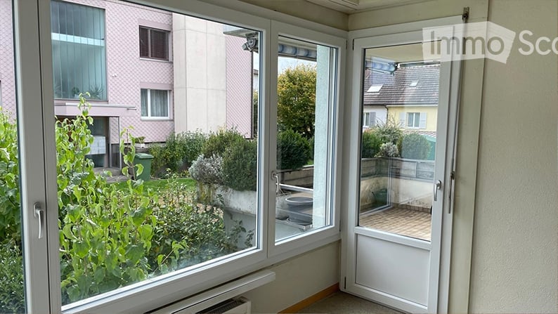 Balcony with glass doors, overlooking a garden area with greenery and neighboring buildings.