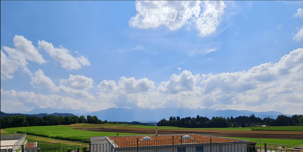 A scenic view of a rural landscape with green fields, scattered trees, and distant mountains under a partly cloudy sky.