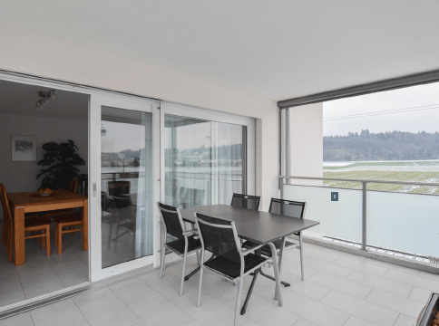 Balcony with large windows, view of water, dining table and chairs inside the room visible through the open sliding doors.