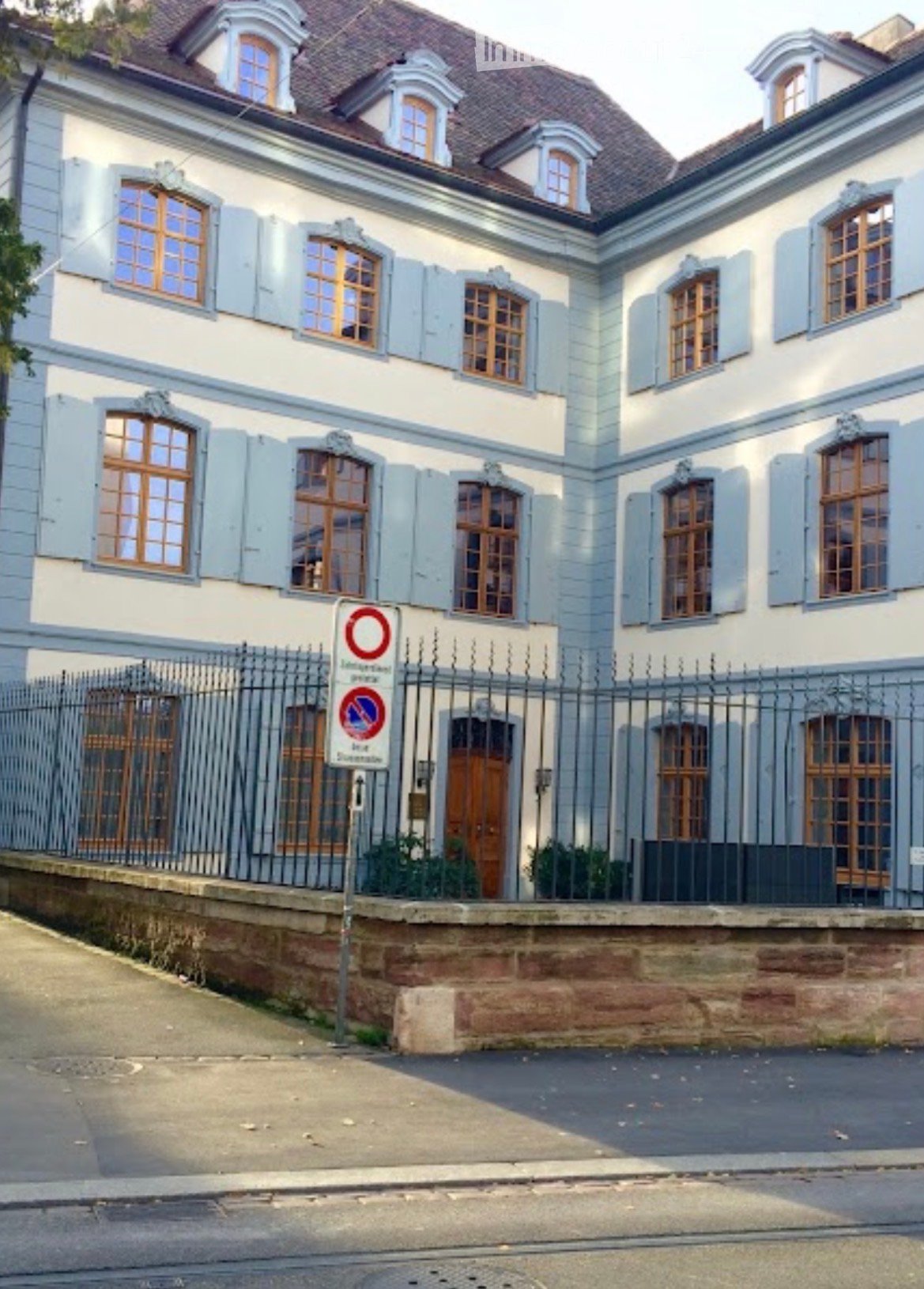 A multi-story residential building with light blue walls, white trim, and multiple windows with wooden frames. The building features a decorative roofline and is surrounded by a metal fence with a raised ground floor visible in front.