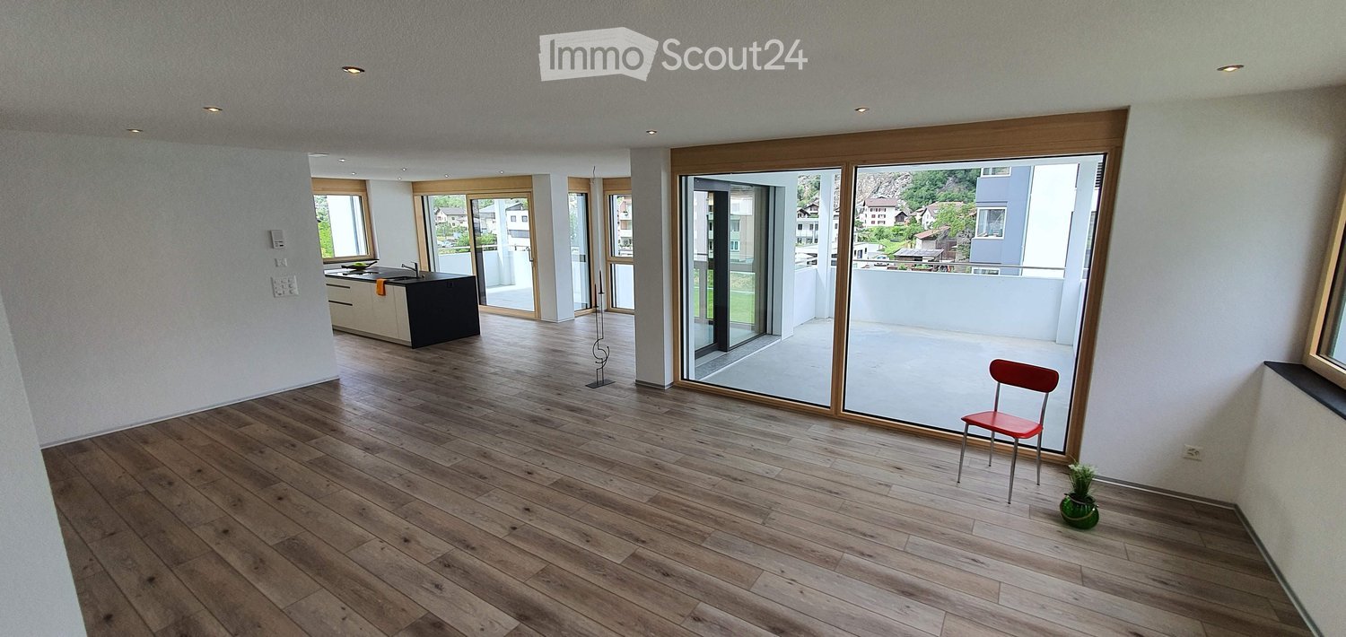 Bright, open living space with wooden flooring, large sliding glass doors leading to a balcony, and a small seating area with a red chair and a potted plant.