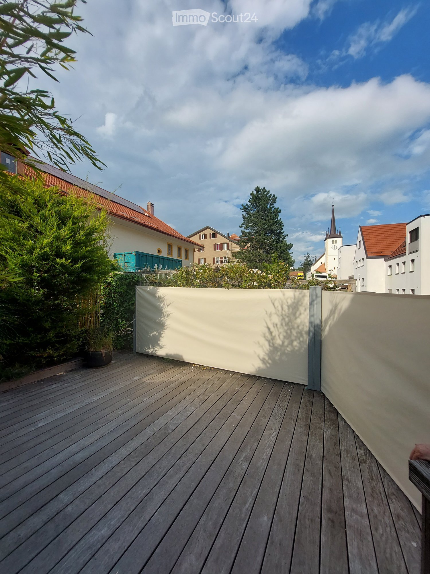 Wooden deck with beige privacy screens, surrounded by greenery and neighboring houses.