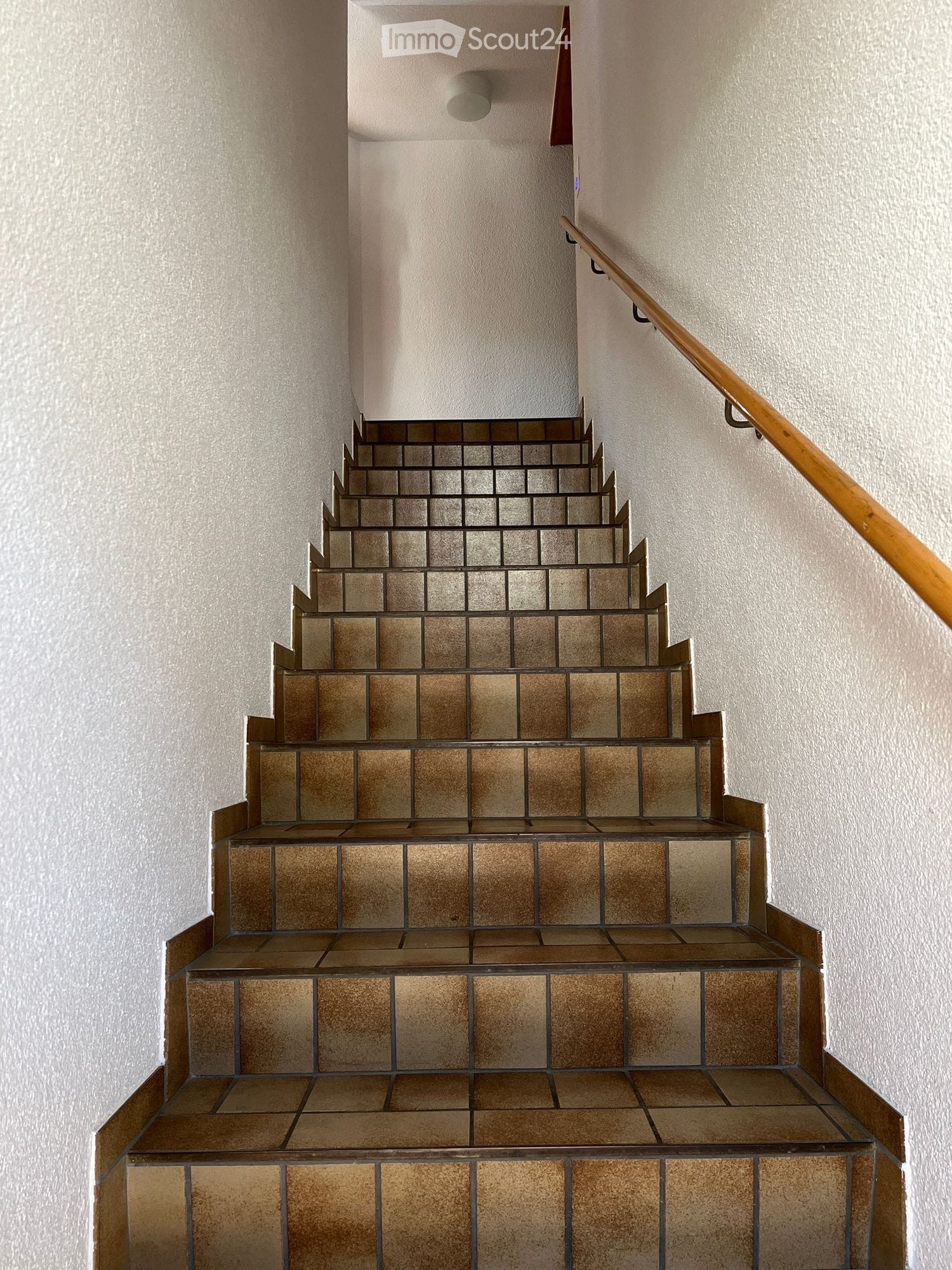 Staircase with beige tiled steps and a wooden handrail, set against light-colored walls.