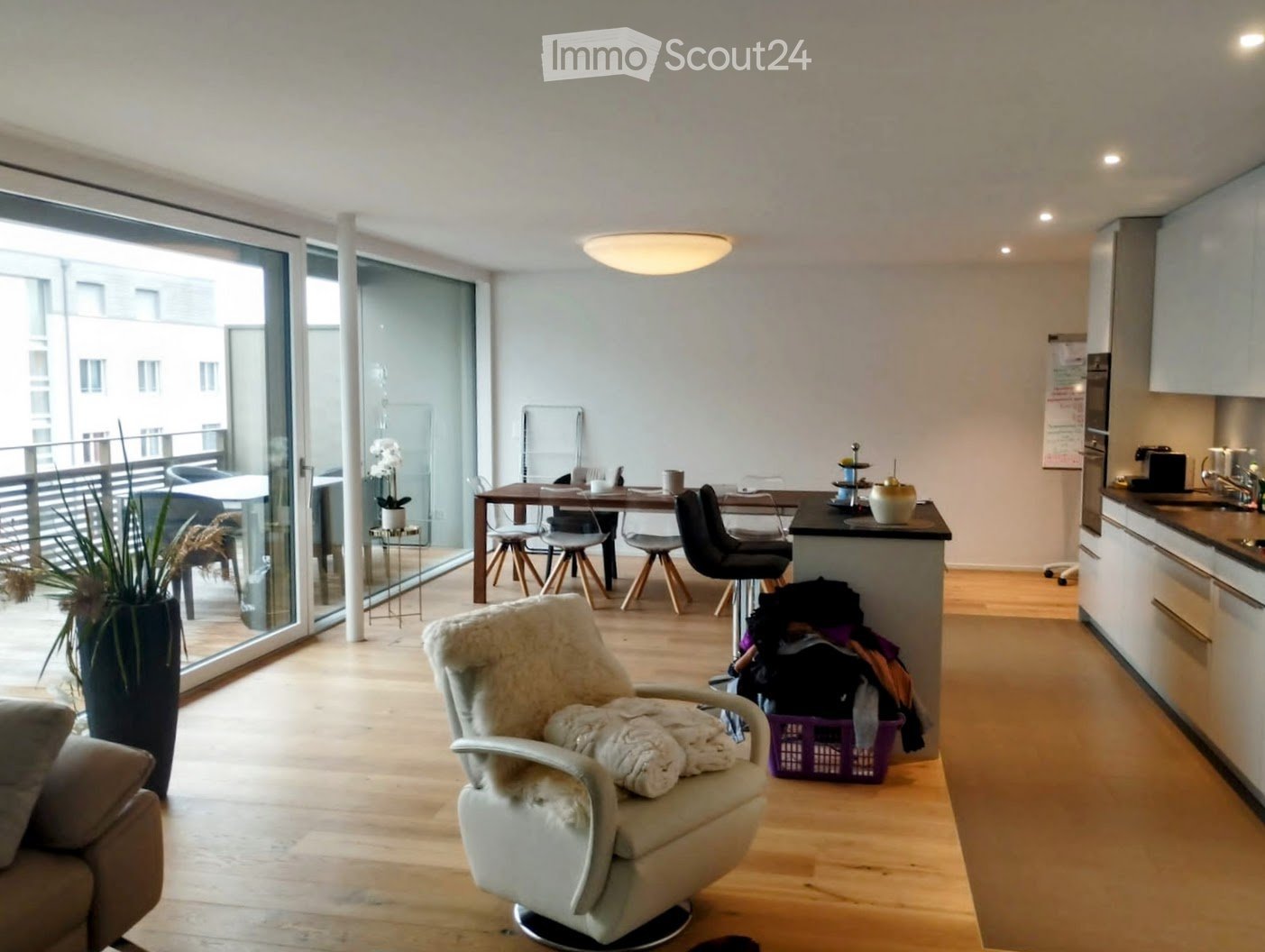 Modern, open-plan living and dining area with light wooden flooring, large windows, and a balcony. Features a white leather armchair, sofa, dining table with chairs, kitchen area with cabinets and appliances, and decorative vases.