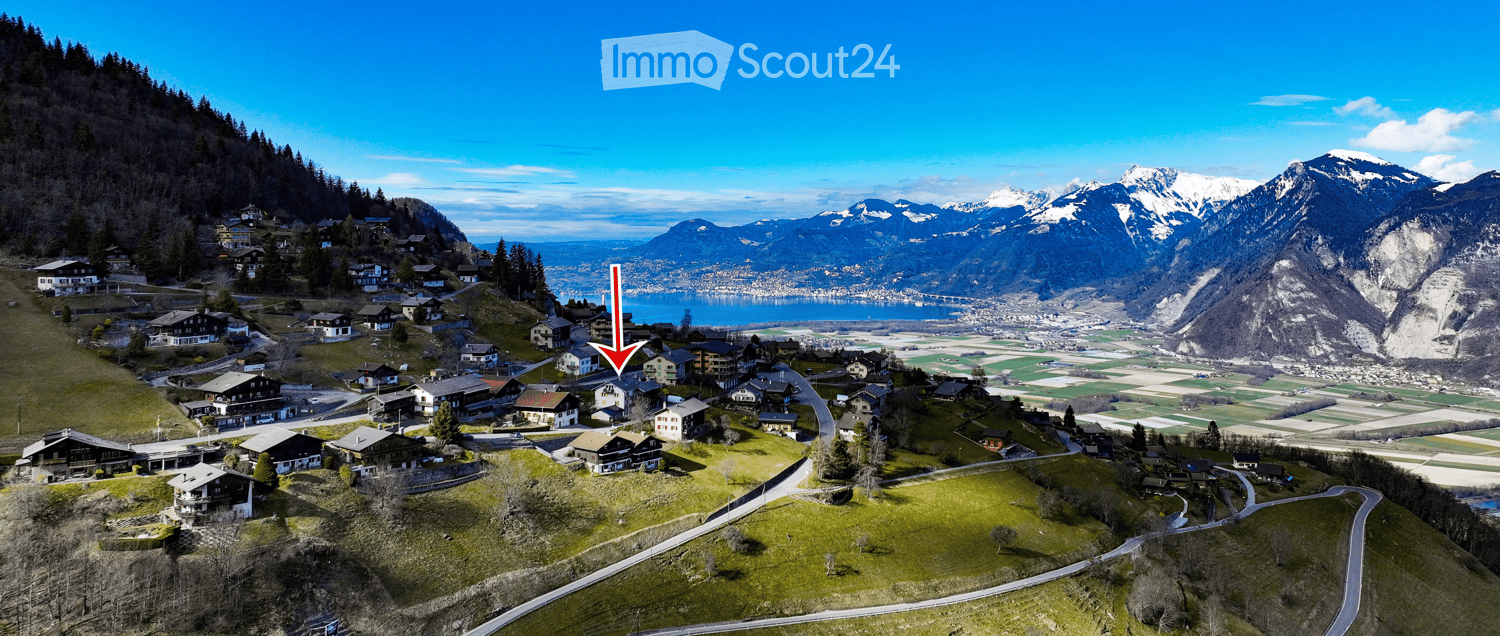Aerial view of a small village nestled in a valley surrounded by mountains and a lake, with scattered houses and winding roads.
