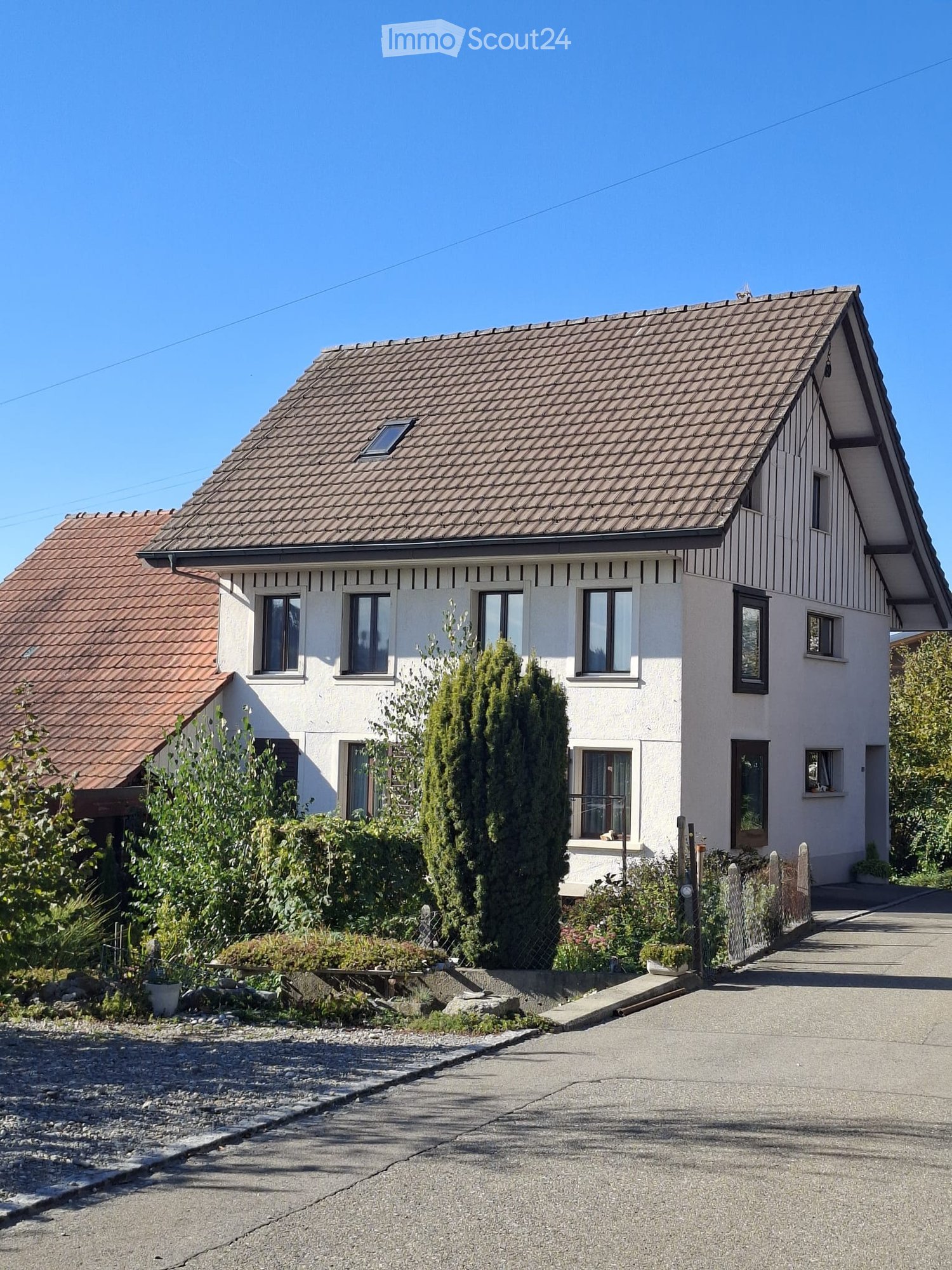 Two-story house with a tiled roof, white exterior walls, multiple windows, and a small garden area with trees and shrubs.