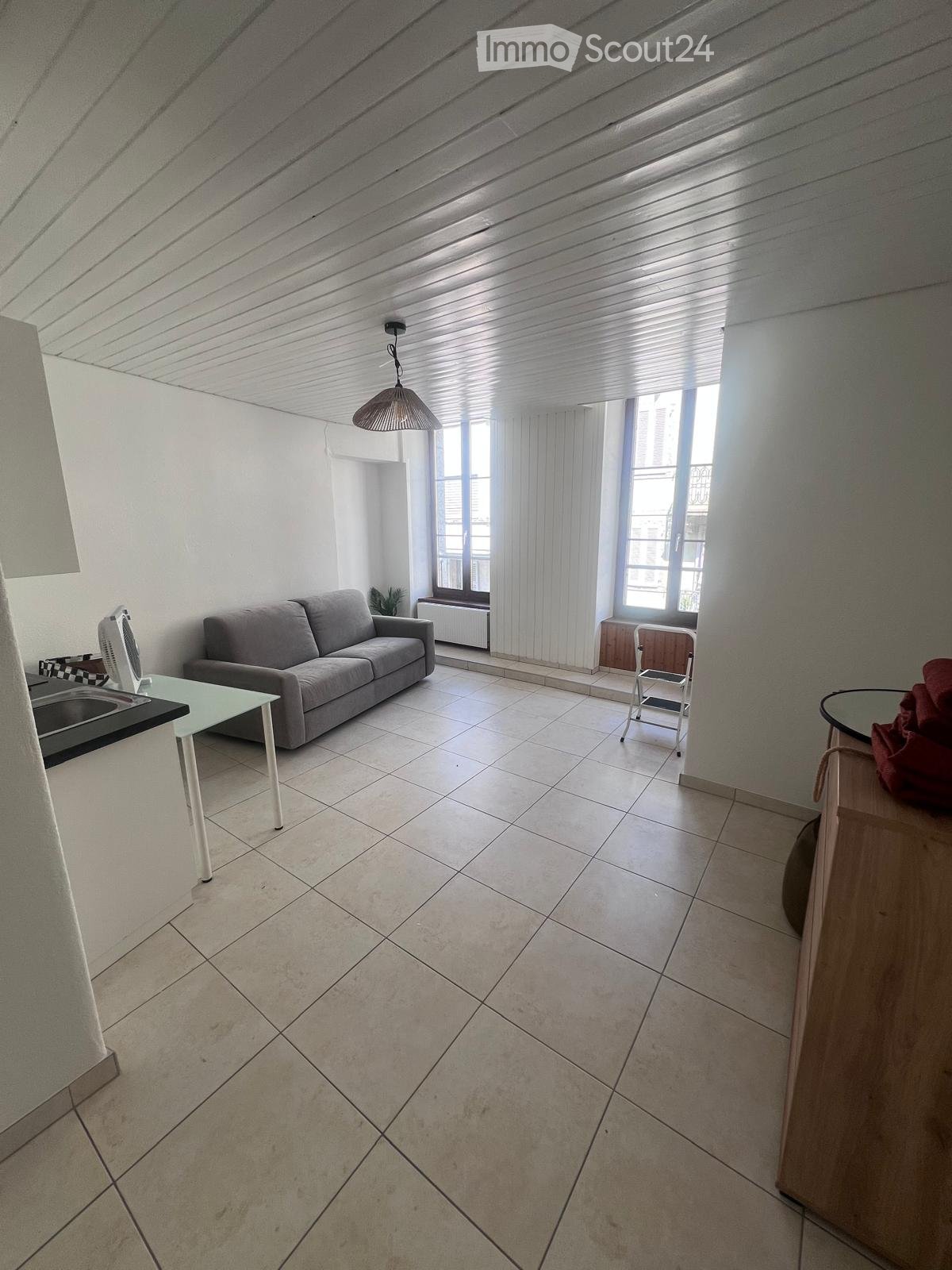 Bright living room with tiled floor, white walls, large windows with curtains, a grey sofa, a glass table, a fan, a small chair, and a mirror on a wooden stand.