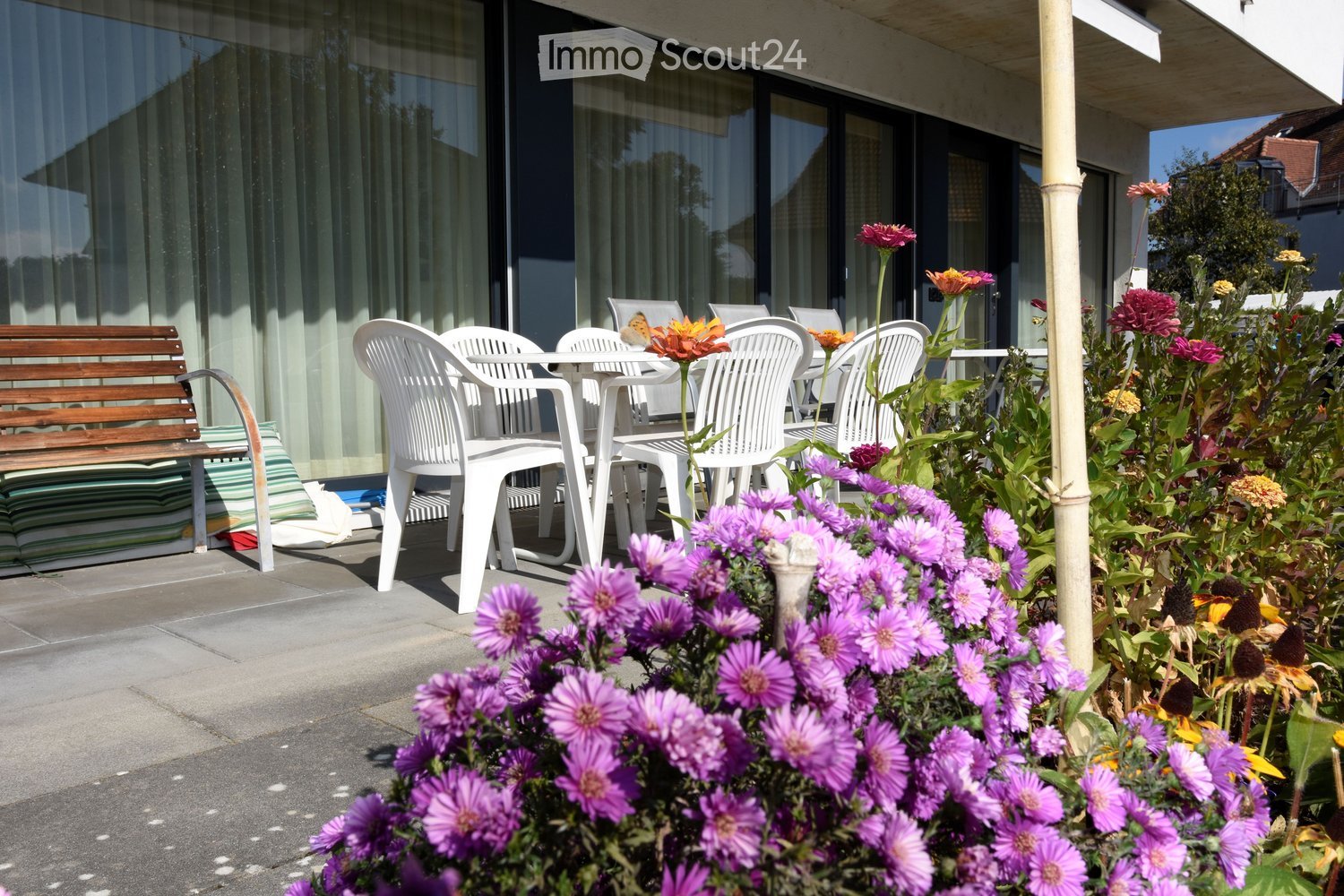 Garden area with a wooden bench, white plastic chairs, and a table surrounded by colorful flowers, near a modern building with large windows.