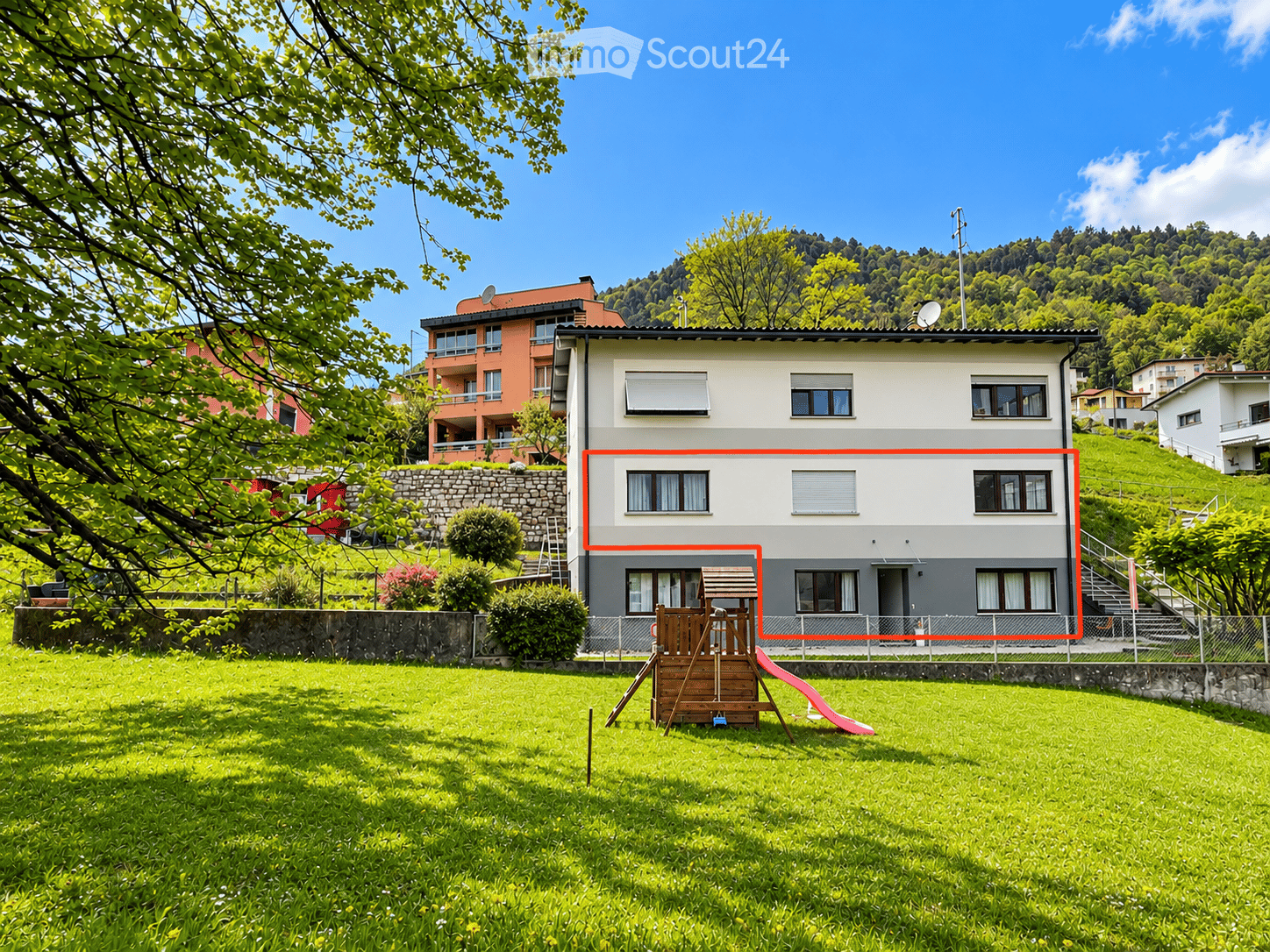 A two-story house with a modern design, featuring large windows and a balcony. The house is surrounded by a well-maintained lawn and trees, with a wooden play structure in the front yard.