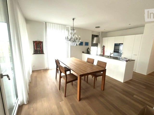 Bright, modern dining area with a wooden dining table and chairs, white kitchen cabinets, a chandelier, and large windows with white curtains.