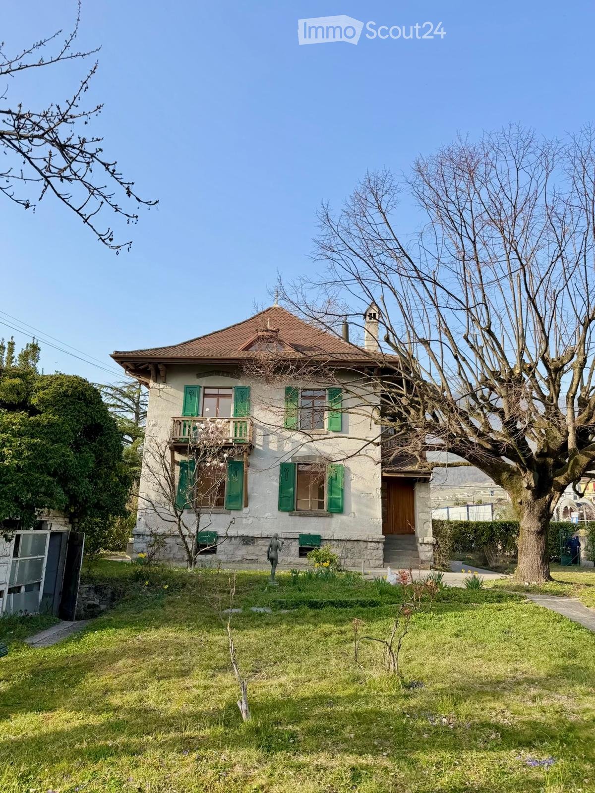 Two-story house with a tiled roof, green shutters, and a balcony on the upper floor. The house is surrounded by a well-maintained lawn and trees.