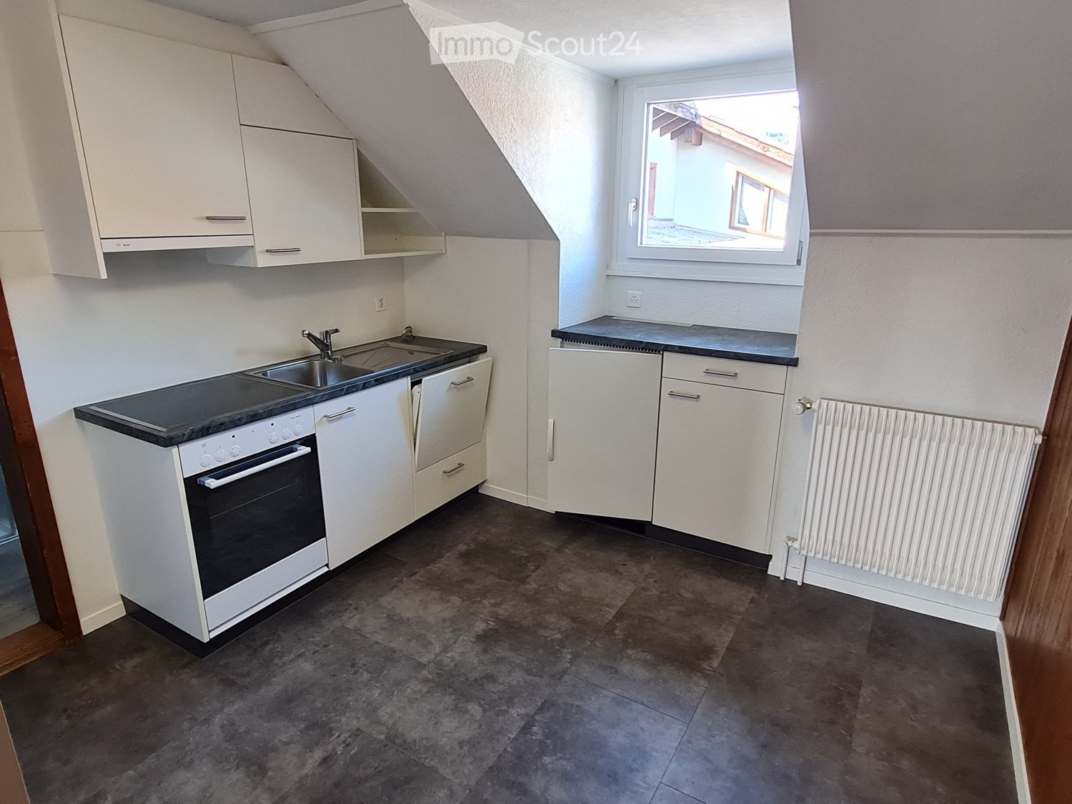 Modern kitchen with white cabinets, dark countertops, sink, oven, and radiator. Features a large window with a view of the outside.