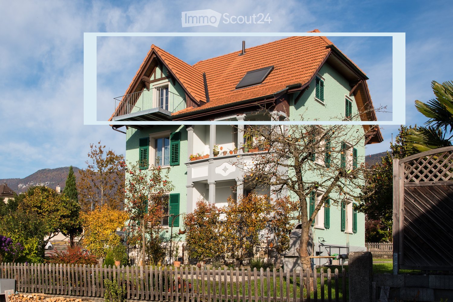 Two-story house with a red-tiled roof, green shutters, and a balcony on the upper floor. The house is surrounded by a wooden fence and various trees and shrubs.
