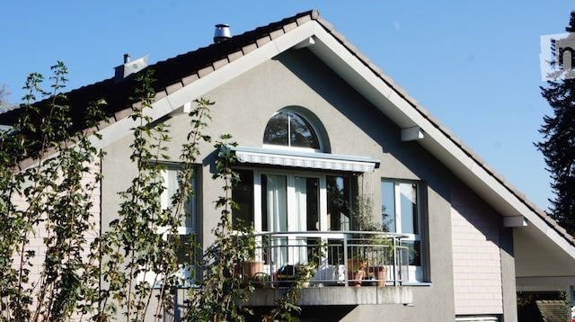 Modern two-story house with a grey exterior, large windows, and a balcony with potted plants.