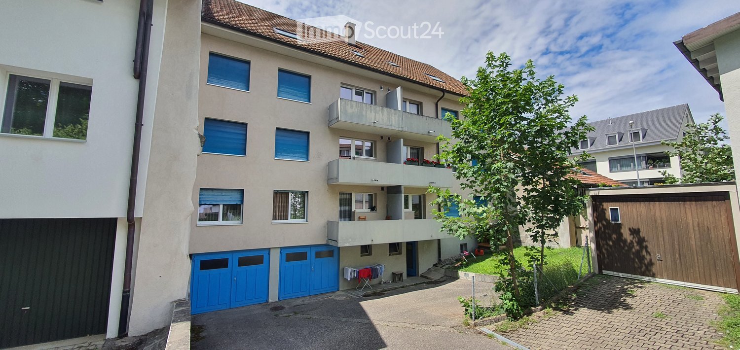 Multi-story residential building with balconies, blue garage doors, and a paved driveway.