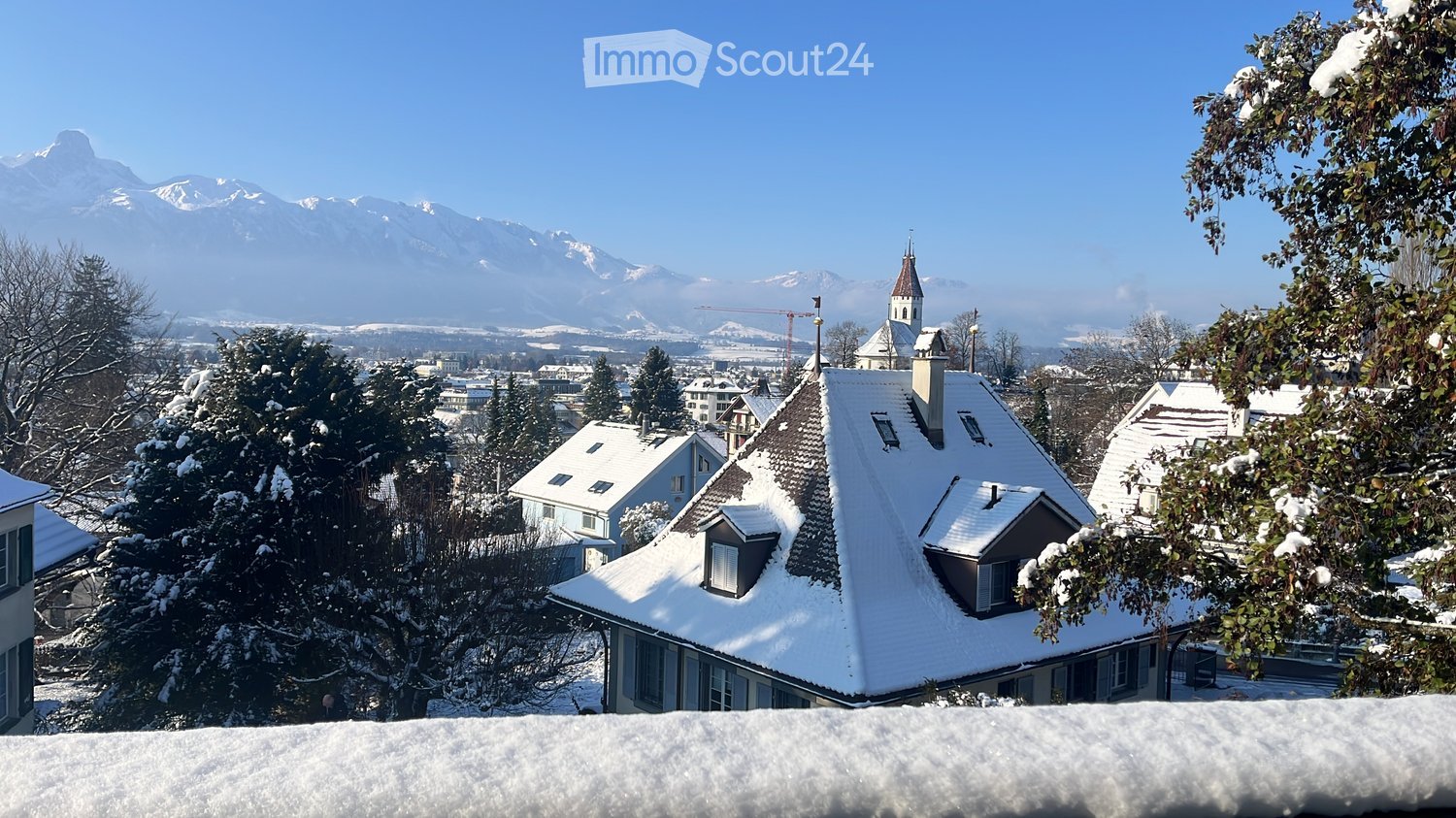 A picturesque view of a snow-covered village with traditional houses and a church tower, surrounded by snow-laden trees and distant snow-capped mountains under a clear blue sky.