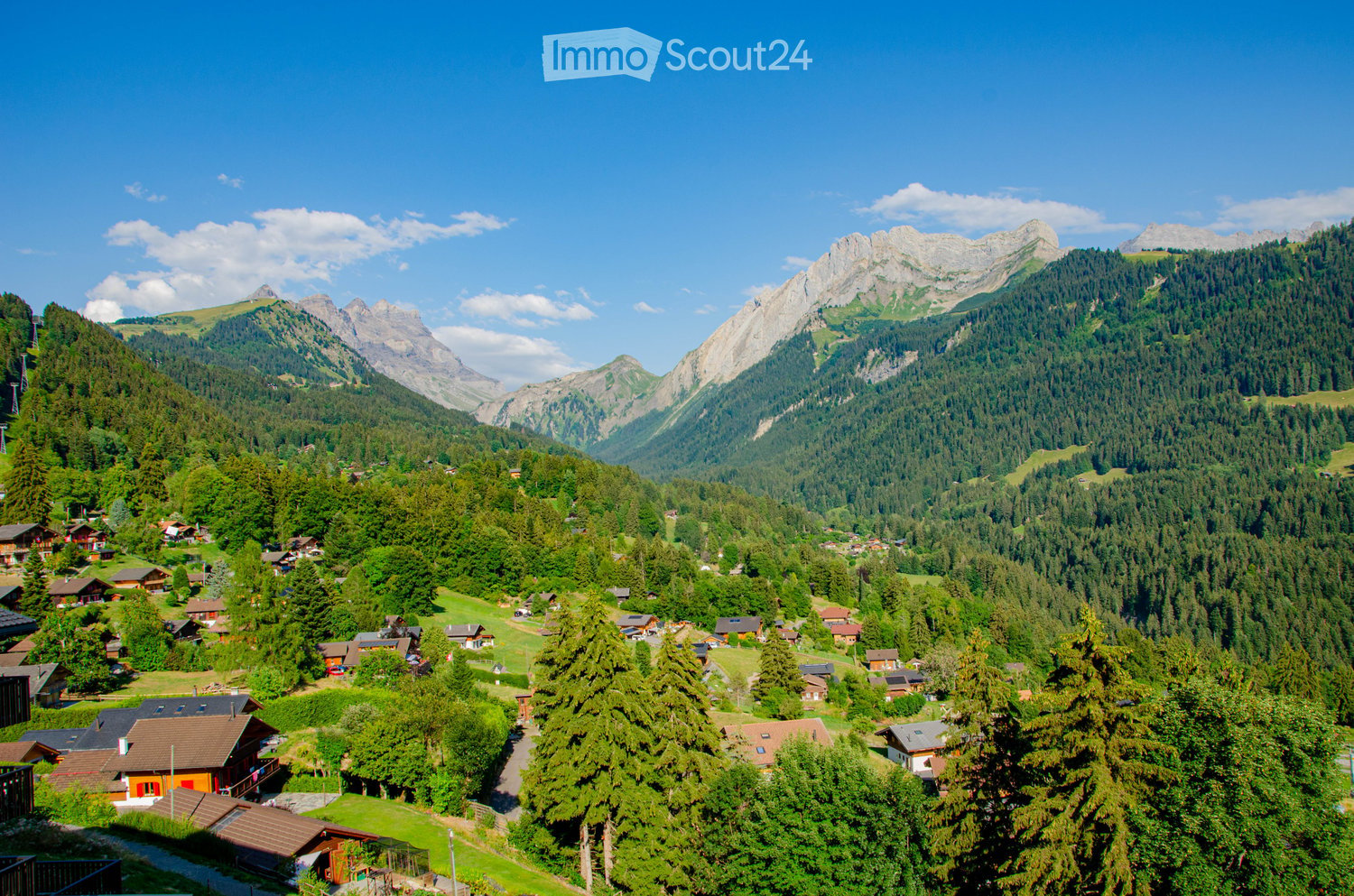 A picturesque alpine village nestled in a lush green valley surrounded by towering mountains. The village features scattered wooden houses with red roofs, surrounded by dense forests and open meadows.