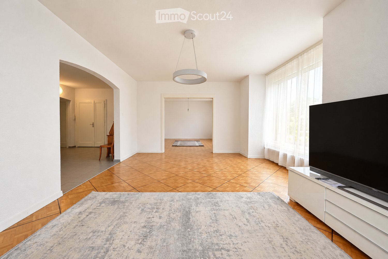 Bright living room with wooden flooring, large windows with white curtains, a modern pendant light, a flat-screen TV on a white stand, a grey rug, and a small wooden chair in the adjacent area.