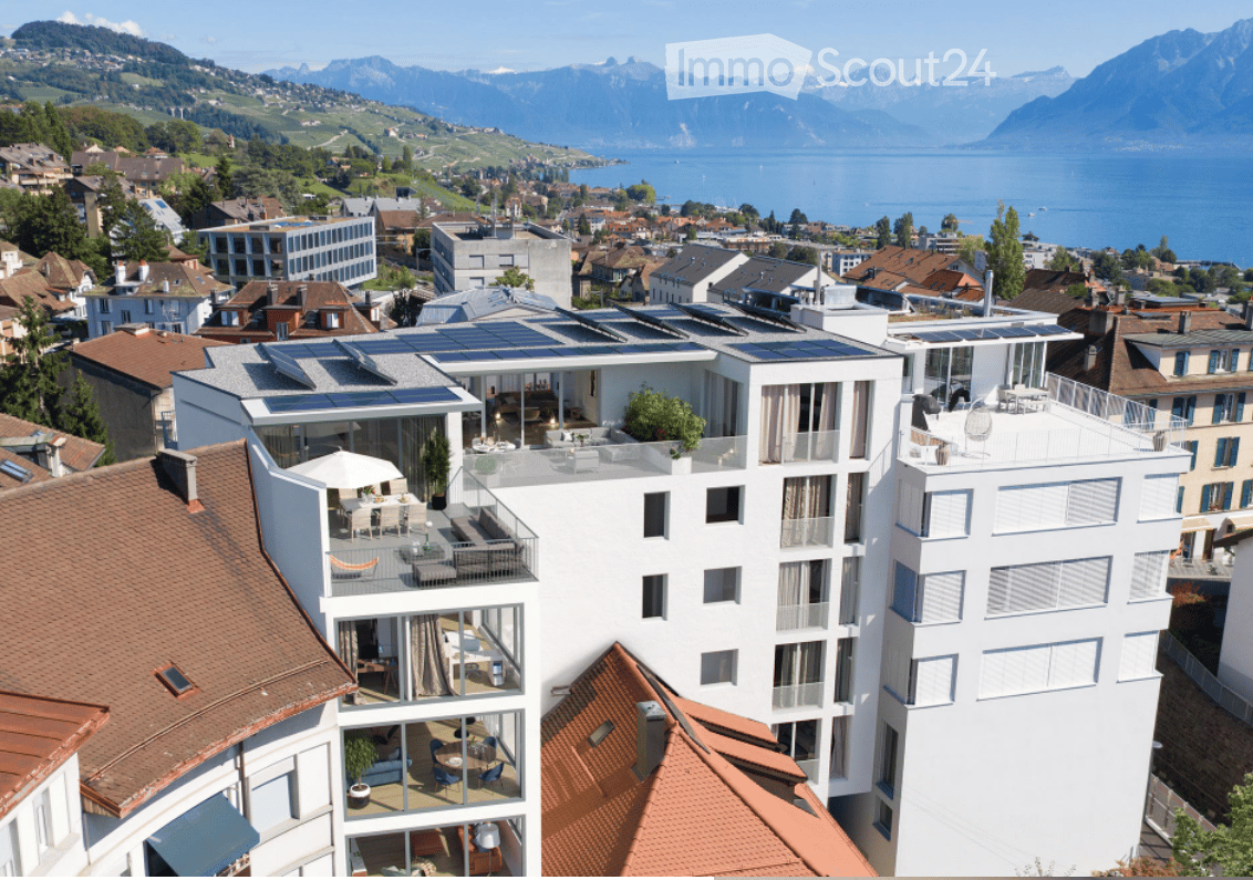 Modern multi-story building with large windows, balconies, and rooftop terraces overlooking a scenic lake and mountain backdrop.