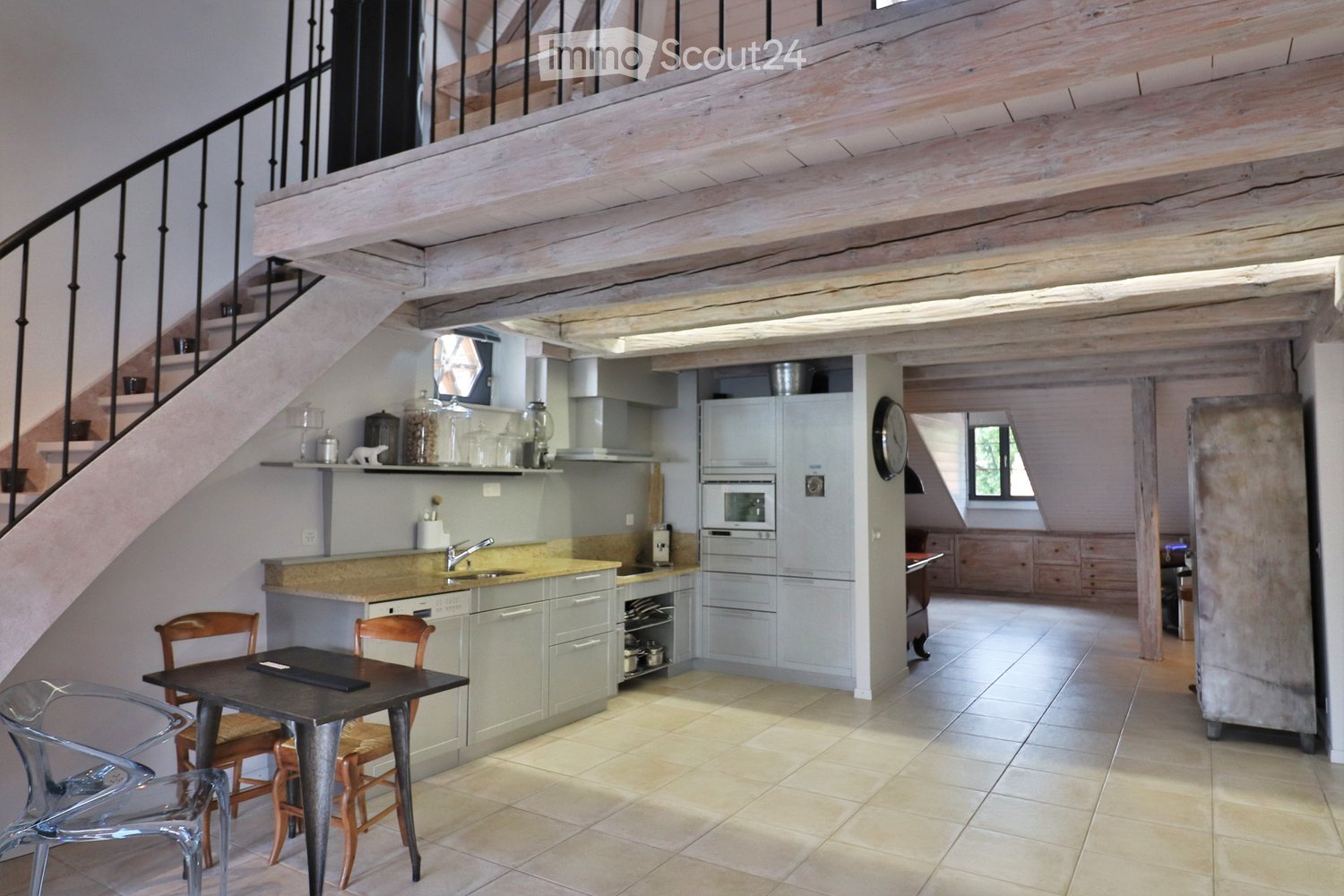 Open-plan kitchen with light grey cabinets, a central island, and a dining area. Features include a sink, refrigerator, oven, and a small dining table with chairs.