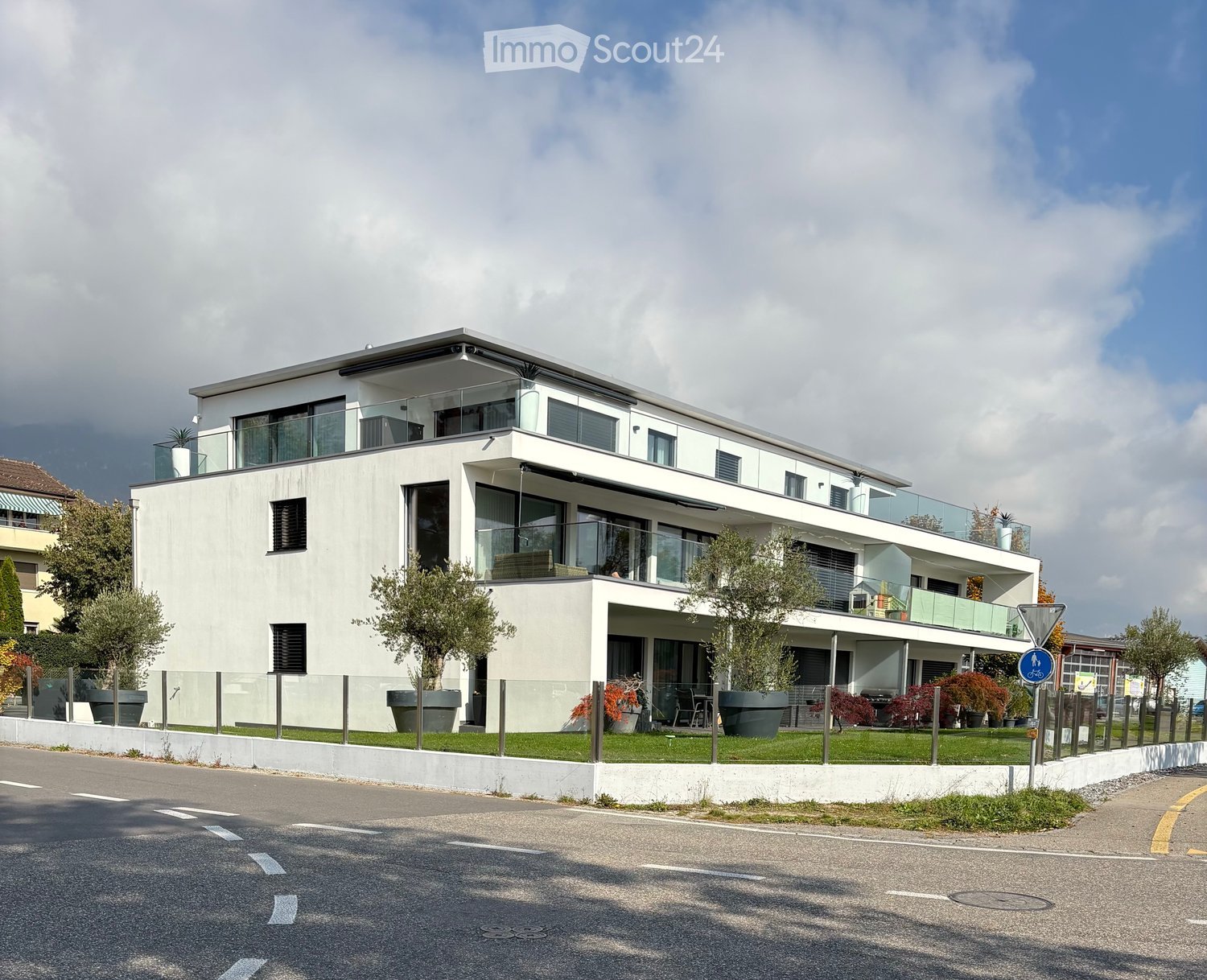 Modern two-story house with white facade, large windows, and balconies, surrounded by greenery and a paved road.