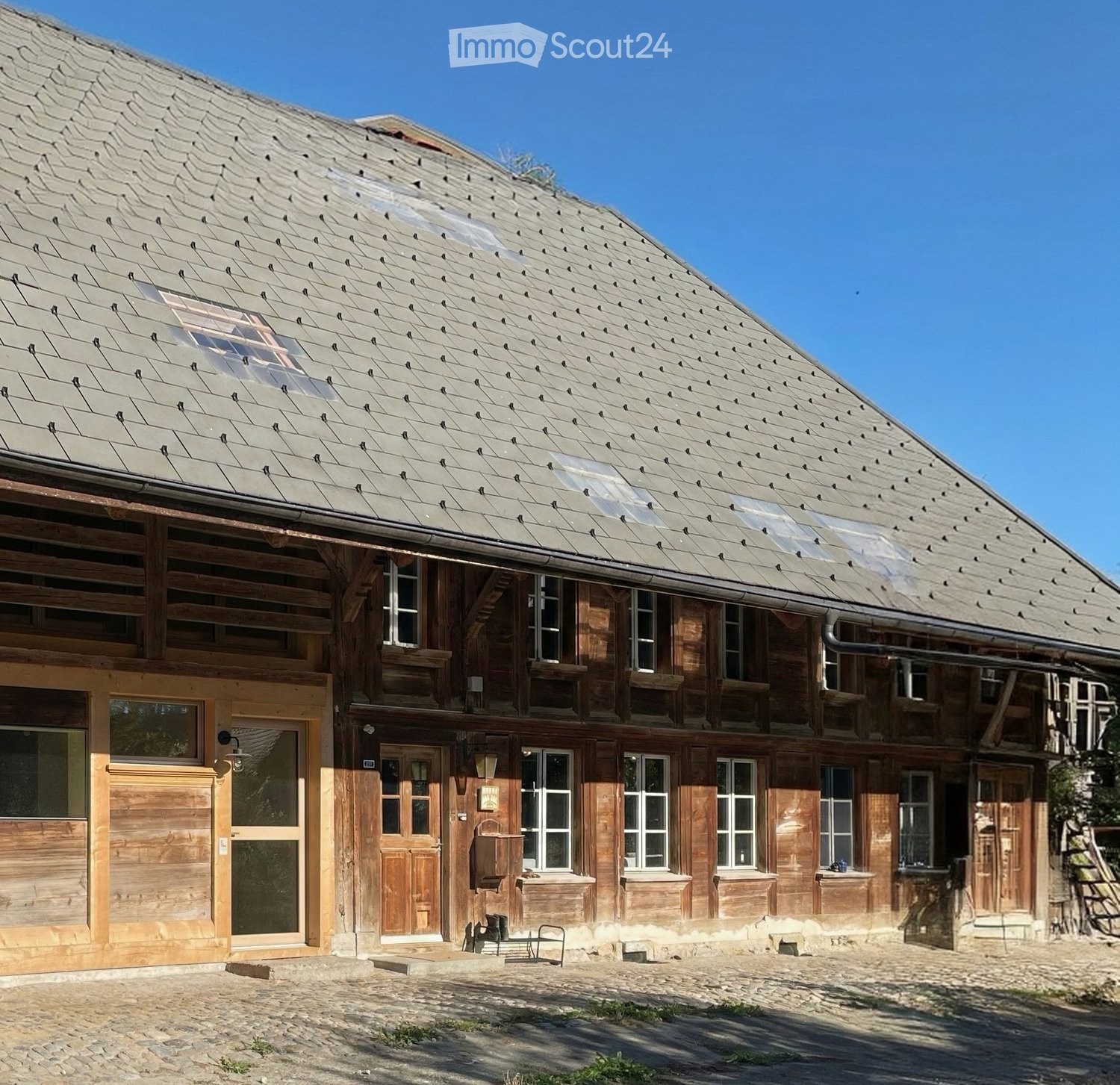 Traditional wooden house with a steep, shingled roof and multiple windows, featuring a stone-paved courtyard.