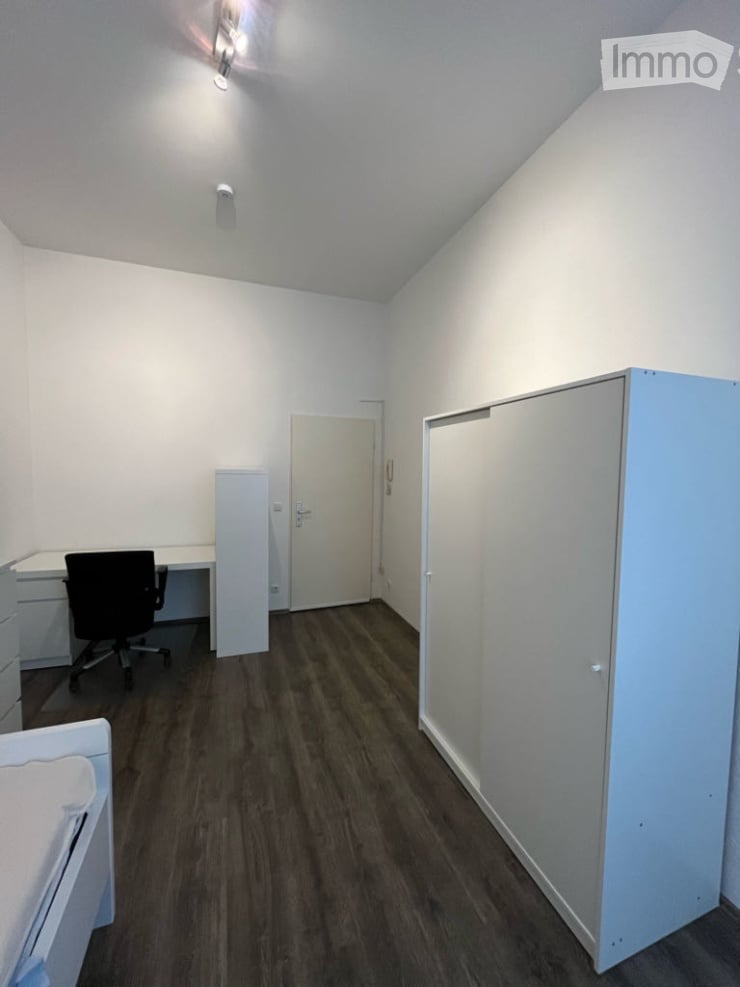 A small, furnished room with a wooden floor, white walls, and a slanted ceiling. It features a desk with a chair, a tall cabinet, and a door leading to another room.