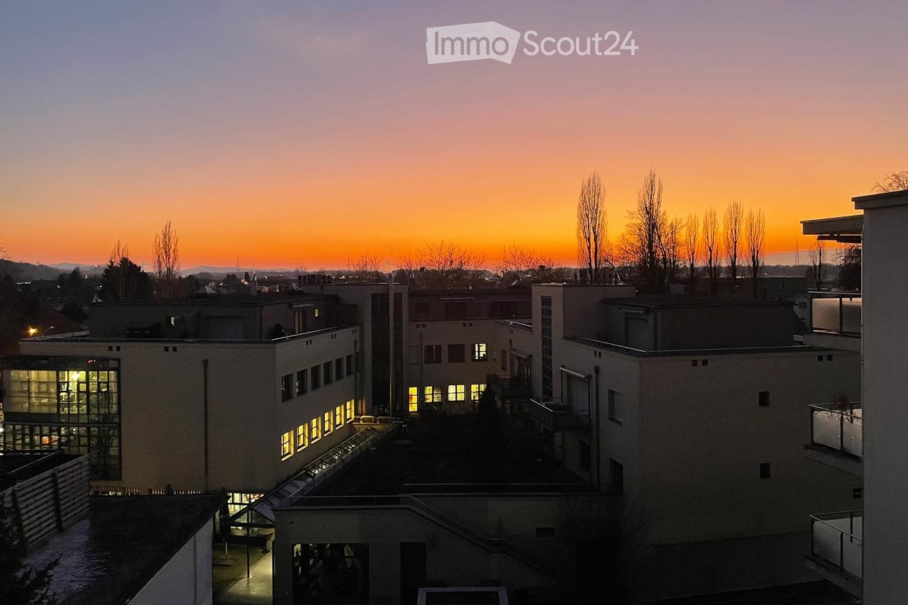 A multi-story building complex with illuminated windows, surrounded by trees and a cityscape at sunset.