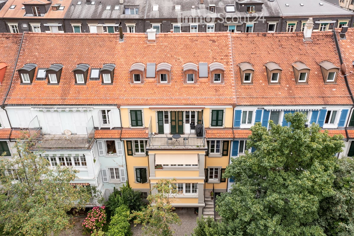 A row of colorful residential buildings with red-tiled roofs, multiple windows, and small balconies. The architecture features varied facades in shades of yellow, blue, and white, with greenery visible in the foreground.
