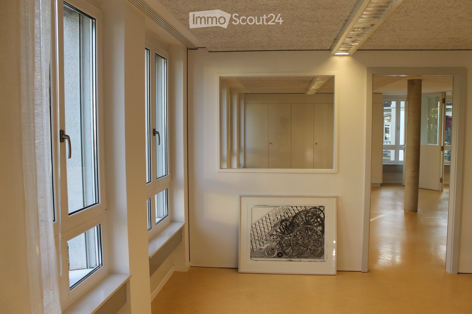 Clean, modern hallway with white walls, large windows, a framed artwork, and a mirror. The floor is light-colored and polished, with a central column visible in the distance.