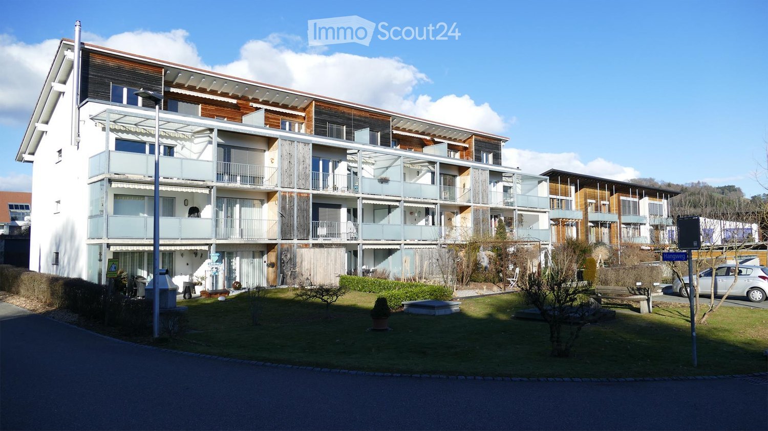 Modern multi-story residential building with balconies, surrounded by a landscaped garden area with benches and potted plants.