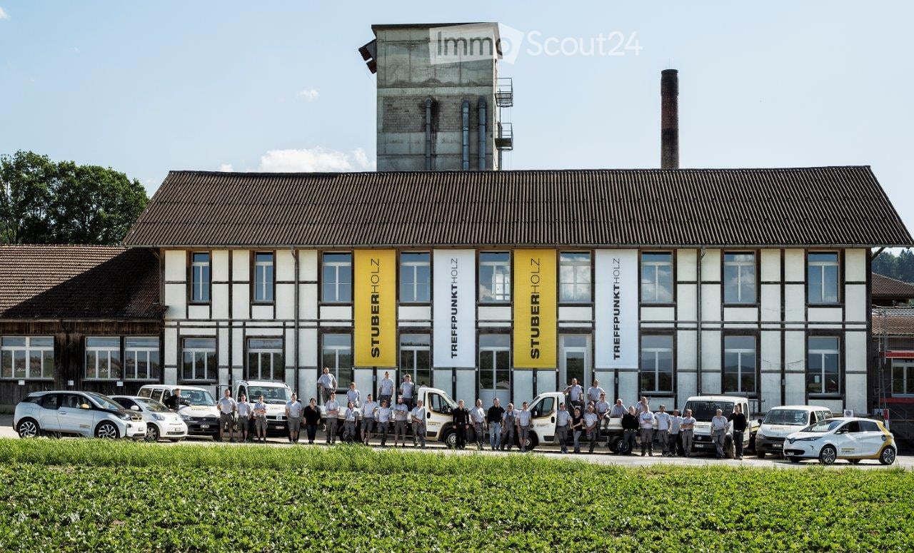 Modern industrial-style building with large windows and signage, surrounded by a group of people and parked cars.