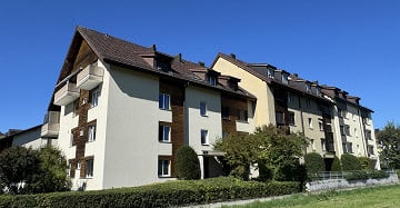 Multi-story residential building with beige and brown facade, multiple windows, balconies, and surrounding greenery.