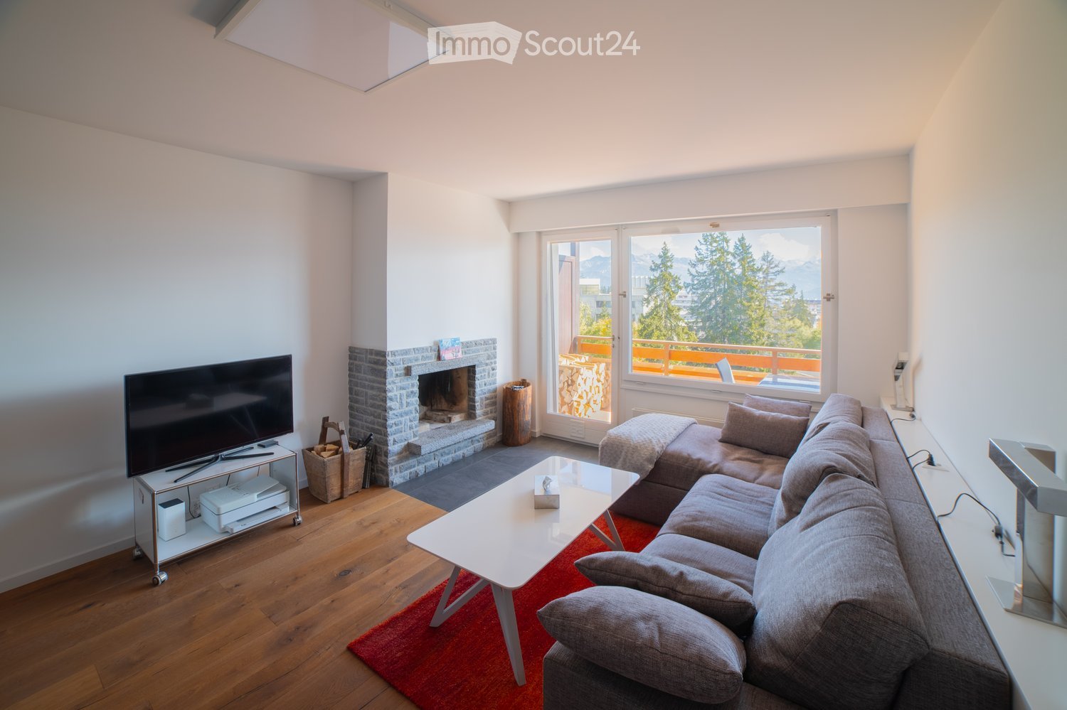 Modern living room with a grey sectional sofa, white coffee table, flat-screen TV, fireplace, and large windows offering a scenic mountain view.
