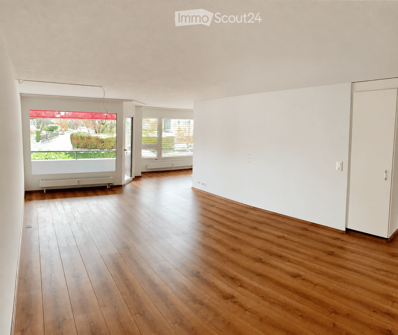 Empty room with wooden flooring, white walls, large windows, and a radiator visible under one window.