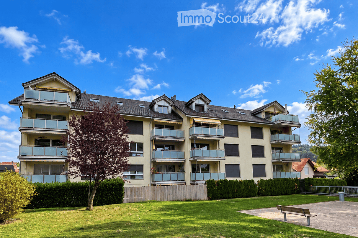 Multi-story residential building with multiple balconies, surrounded by greenery and a small park area with a bench.