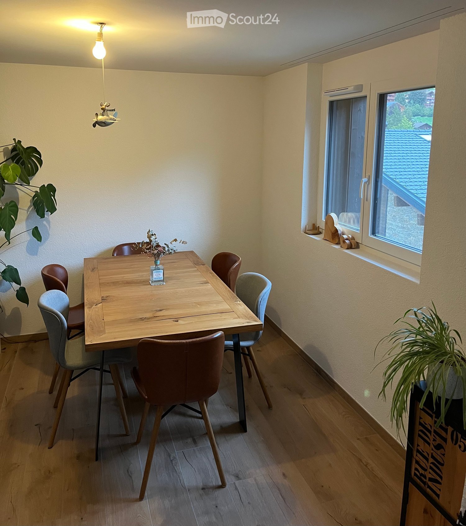 Dining area with a wooden table, six chairs, a vase with dried flowers, and potted plants near the windows.