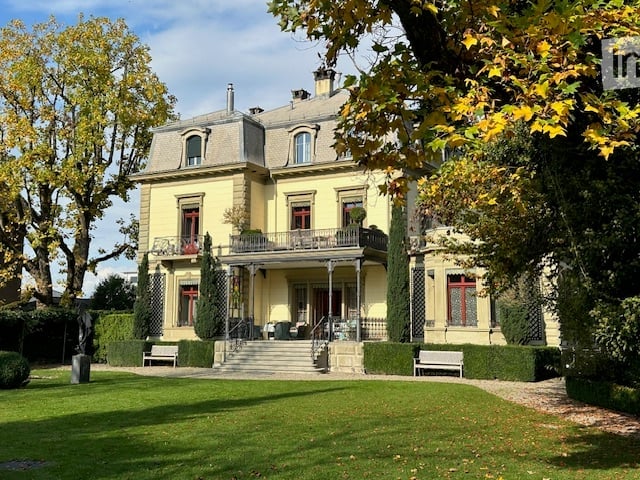 A large, elegant yellow mansion with multiple stories, grey roof, and ornate architectural details. The house features a balcony, manicured lawn, and surrounding trees.
