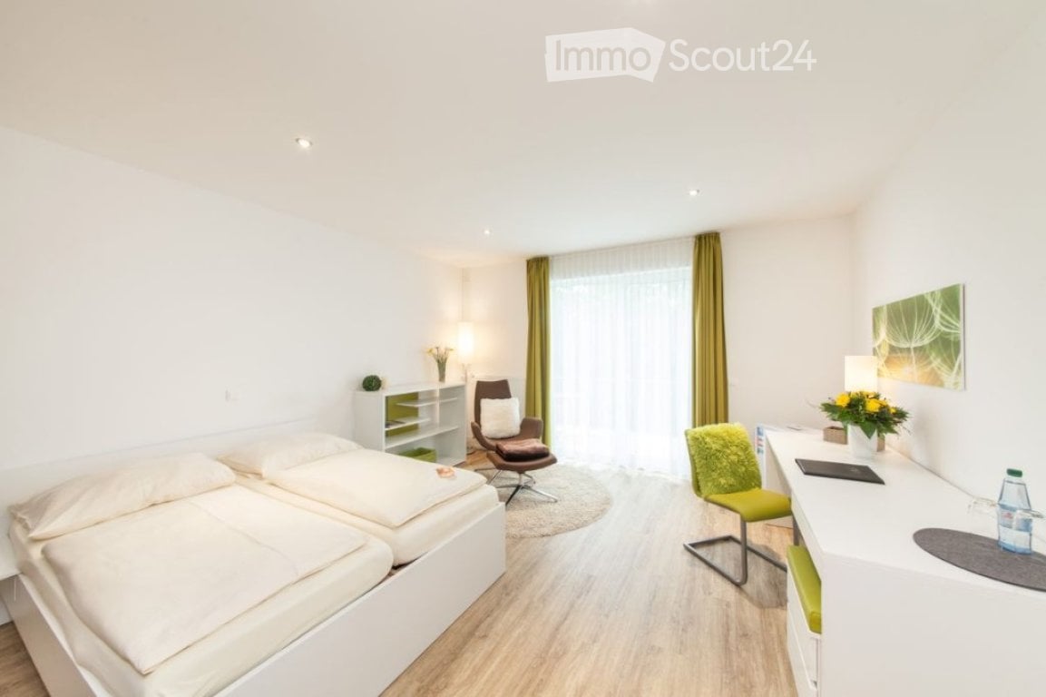Bright, modern bedroom with two single beds, a green accent chair, a desk with a lamp, and a small shelf with decorative items.