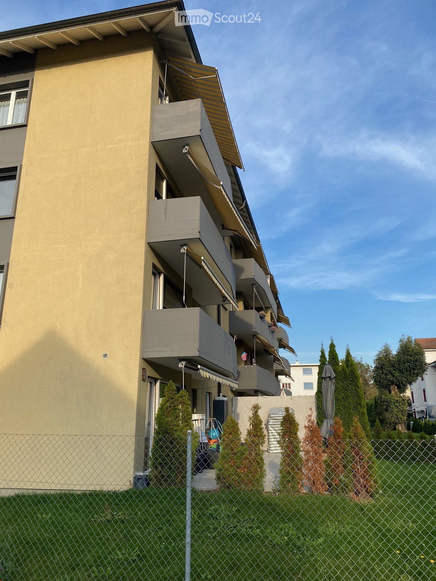 Multi-story residential building with beige facade, multiple balconies, and awnings. The building is surrounded by a fence and small trees.