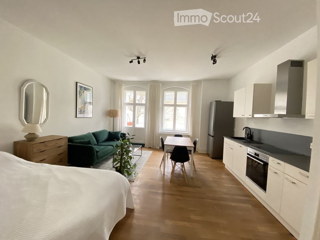 Open-plan living and dining area with a green sofa, dining table with chairs, kitchen area with cabinets, refrigerator, and oven, along with a bed visible in the foreground.